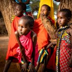 Four young girls in traditional African dress watch on from the base of a large tree.