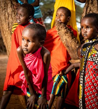 Four young girls in traditional African dress watch on from the base of a large tree.
