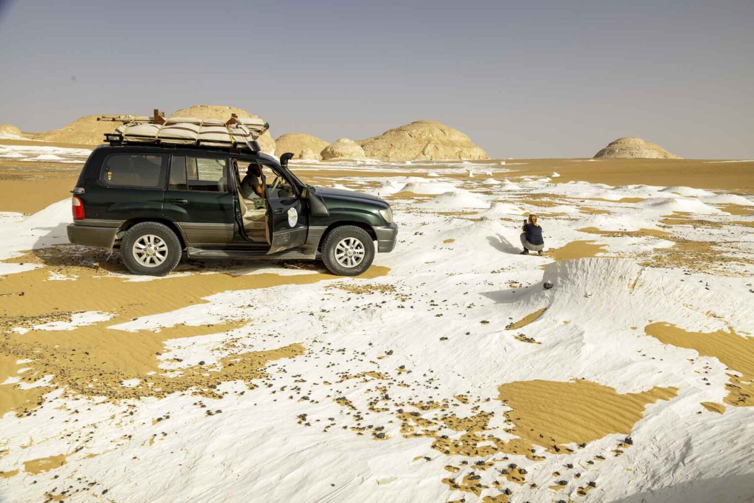 A black four-wheel drive with a door open in the middle of a desert surrounded by low piles of pure white sand. A person crouches in the sand in front of the car.