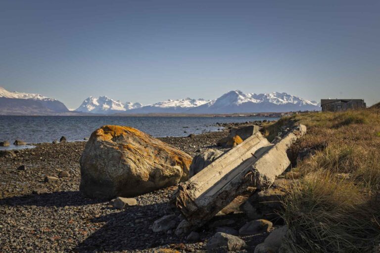 Concrete pillars lie on their side next to a large rock on the stony banks of a lake in front of a snow covered mountain range. The sky is blue.