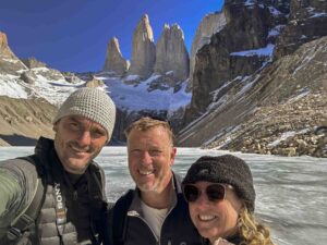 A woman and two men stand in front of frozen lake smiling at the camera. Behind the lake is a rocky mountain range with three distinct narrow peaks.