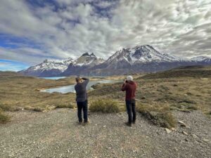 Two men stand with their backs to the camera. They are photographing a lake in front of snow covered mountains